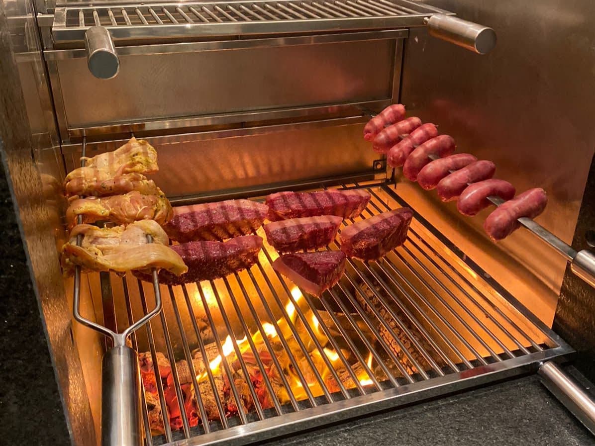 Churrasqueira revestida em inox em uso, com picanha na grelha e espetos de frango e linguiça sobre a brasa
