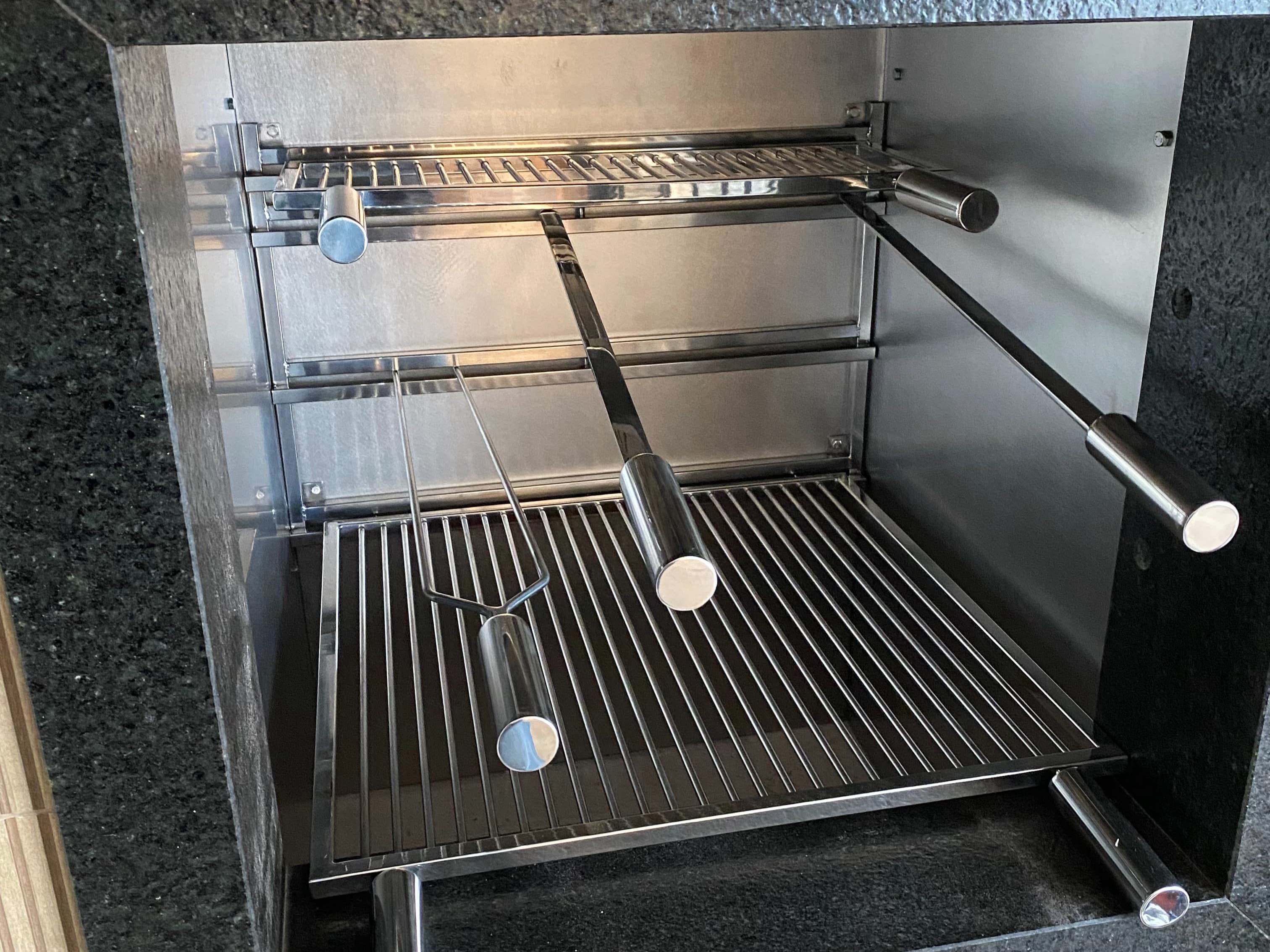 Espetos de inox encaixados no suporte aéreo de uma churrasqueira revestida em inox, sobre a grelha