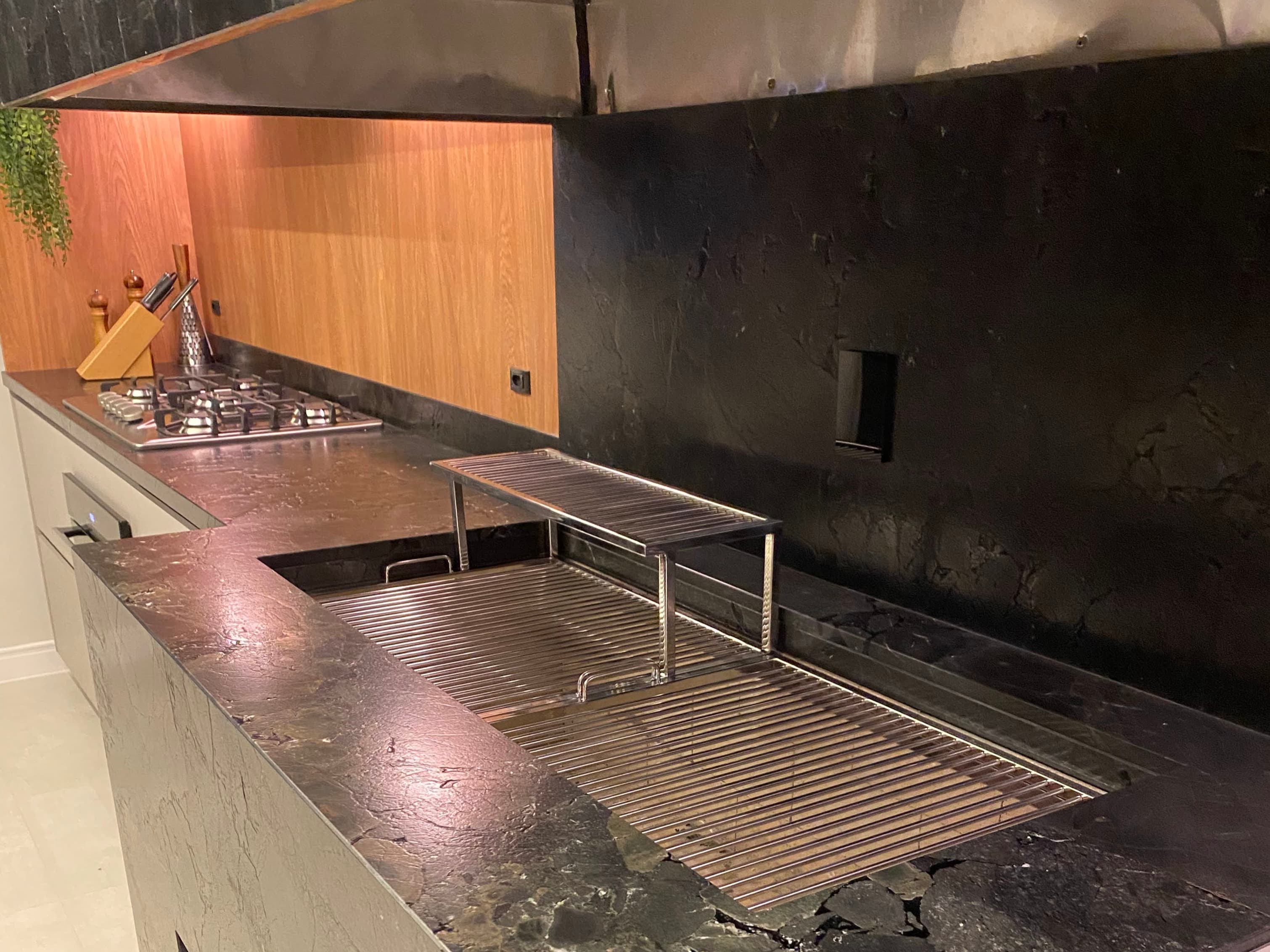 Churrasqueira de inox embutida em bancada de pedra escura, com grelha de altura regulável, em área gourmet