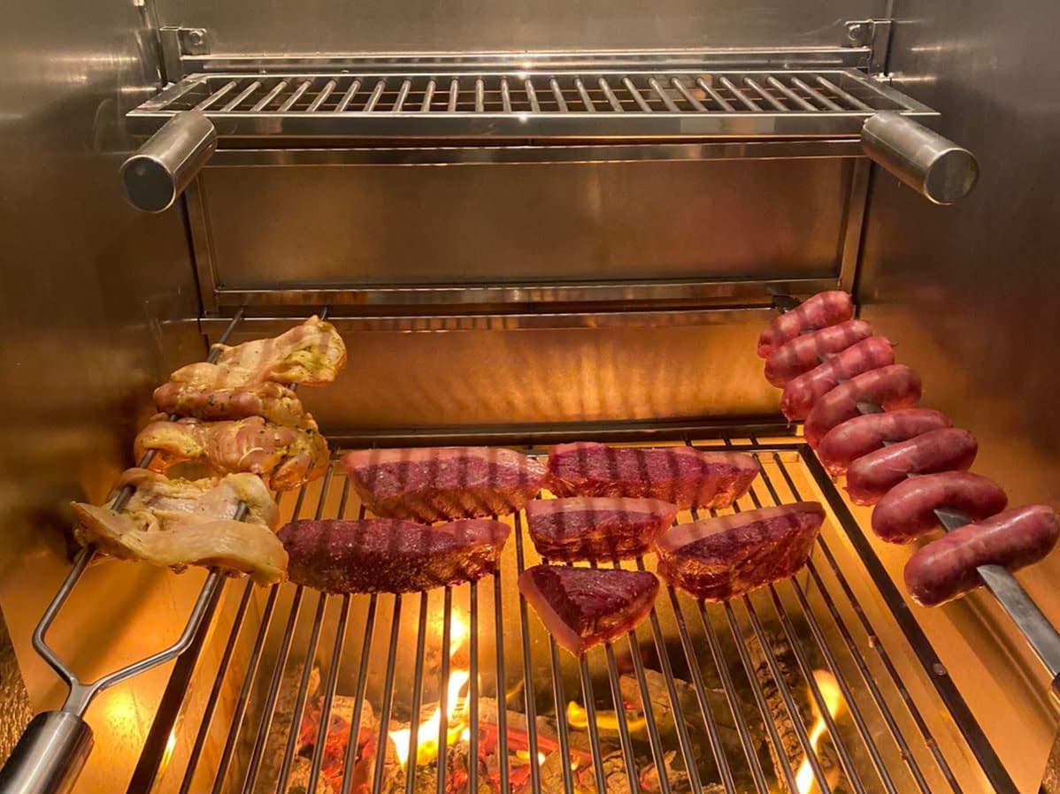 Grelha de inox com carne e linguiça assando sobre a brasa, dentro de churrasqueira de inox